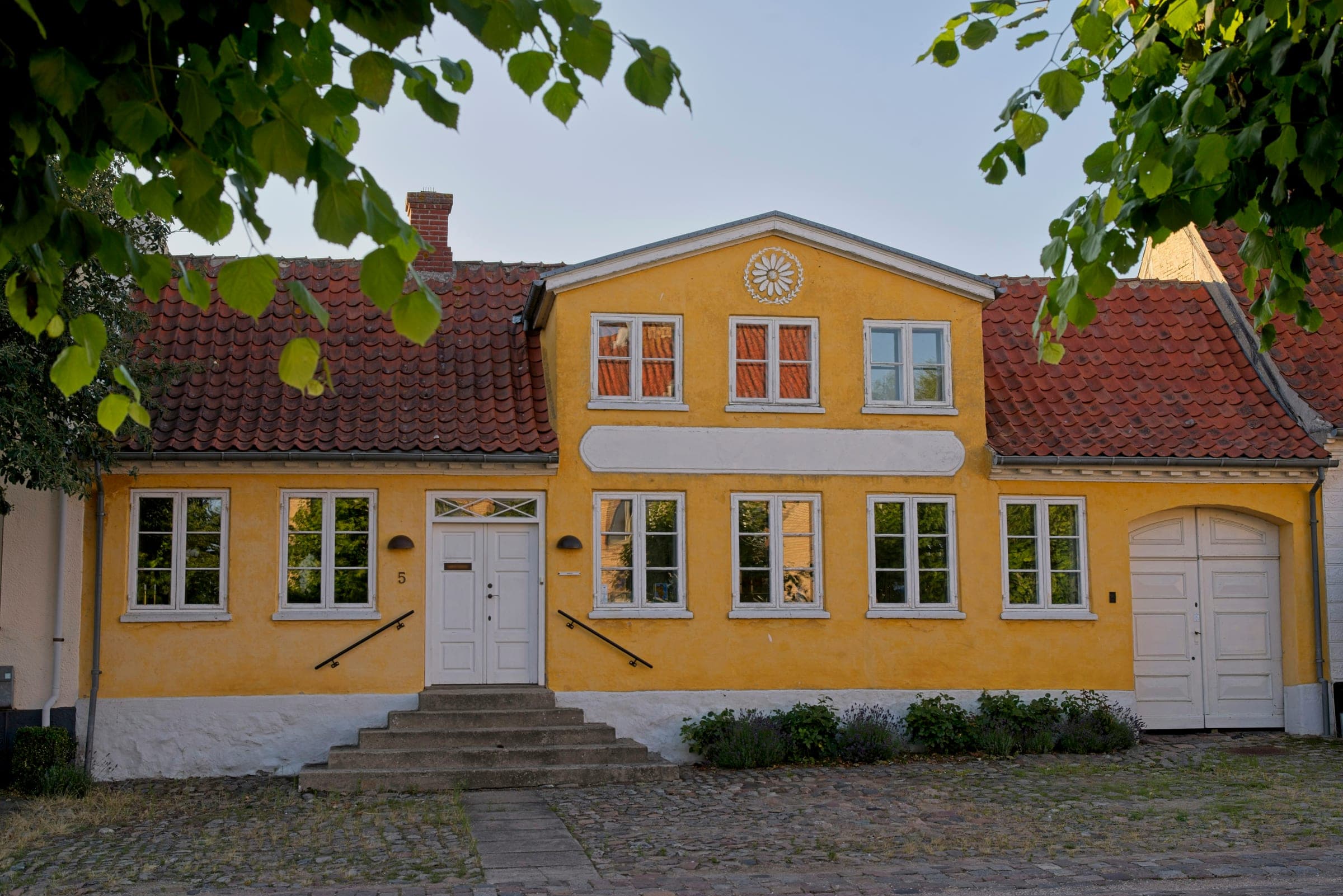 Et dansk provinshus med okkergul facade, rødt tegltag, hvide vinduesrammer og hvid fordør, fotograferet i øjenhøjde i blødt aftenlys uden mennesker eller biler.