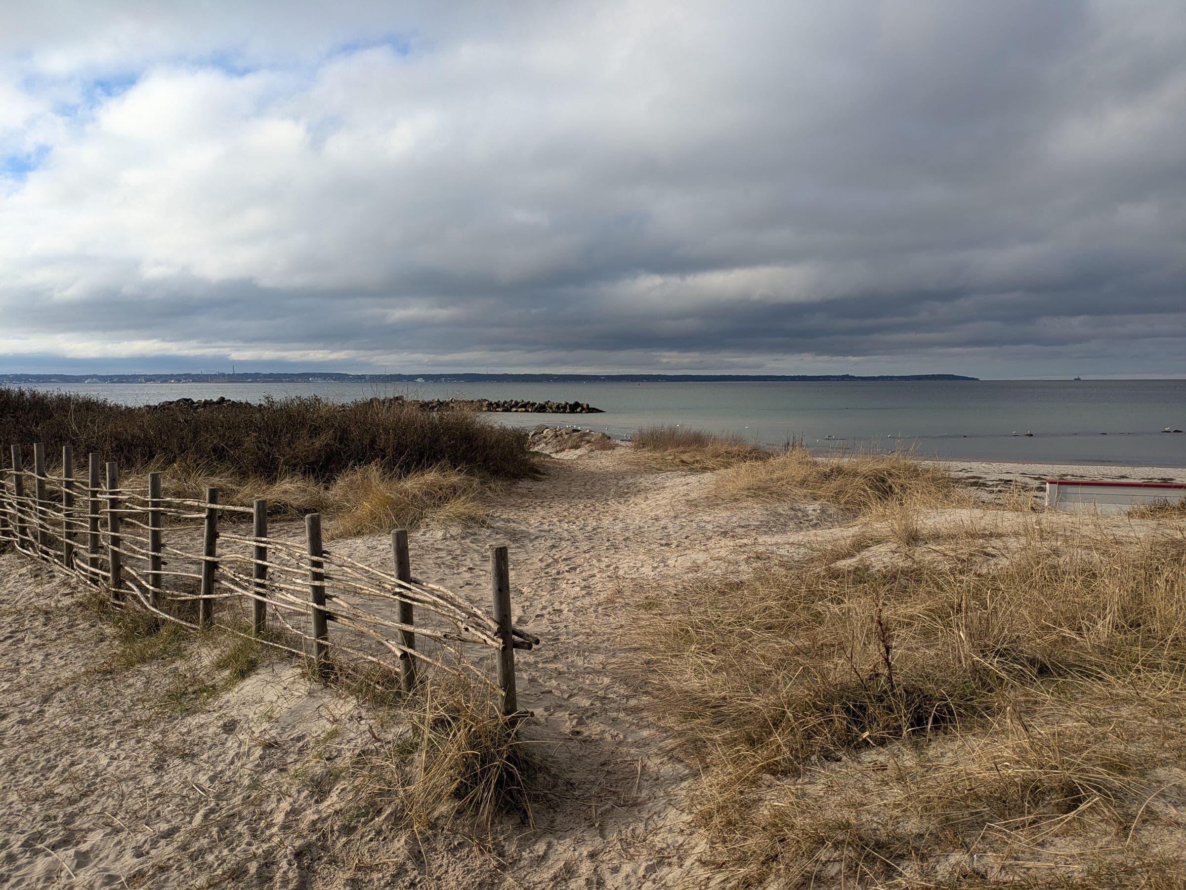 Sandsti gennem klitter med et gammelt flettet trægærde og strandgræs, der fører ned mod en rolig, gråblå kyst under en overskyet himmel.