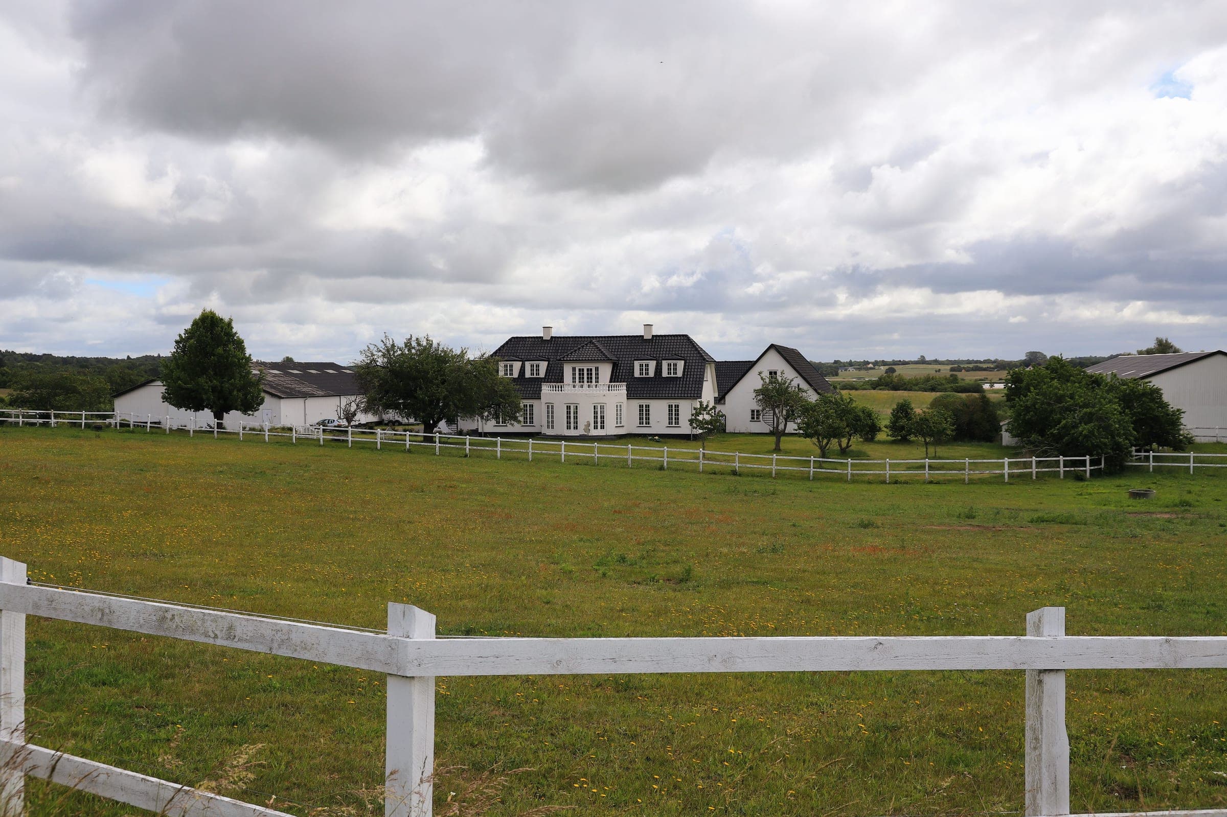 Hvidkalket dansk gård med sort tegltag i et bølget, grønt landskab under en overskyet himmel; et hvidt trægærde ses i forgrunden.