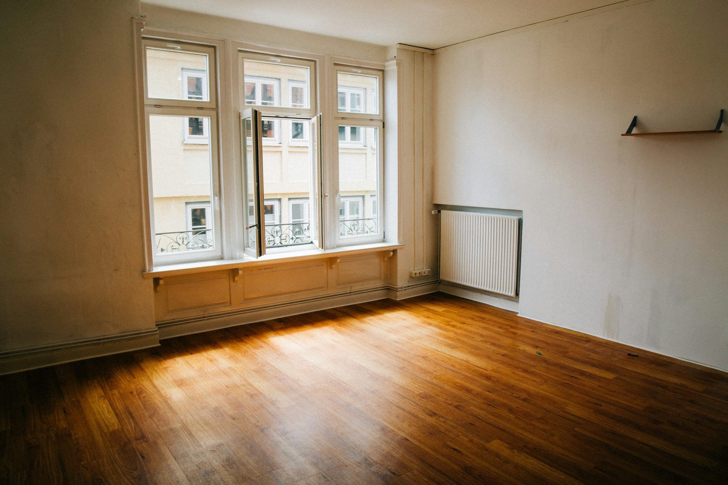Et tomt, nyistandsat værelse med brede vinduer, radiator under vindueskarmen og et varmt trægulv i eftermiddagslys.