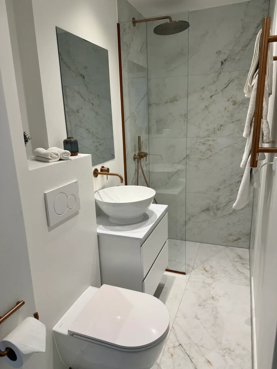 Badeværelset set fra døren: væghængt toilet, håndvask og bruseniche i marmor med messinghåndklædetørrer.
